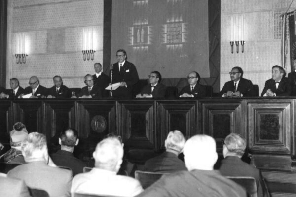 Agostino Origone speaks at the ICTP inauguration ceremony, 1964 A person stands before a large audience and next to other guests of honour. He is Agostino Origone, the rector of the University of Trieste, and he is delivering a speech.