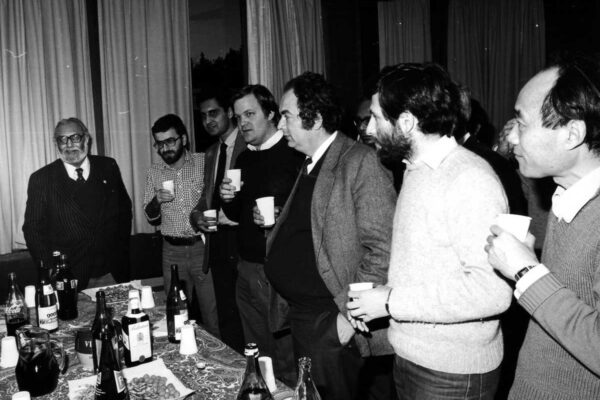 Experimental verification of Salam's electroweak theory, 1983 Scientists L. Bertocchi, G.C. Ghirardi, H.R. Dalafi, L. Fonda and Abdus Salam celebrating together. They stand in a meeting room, surrounding a table with wine and soda. They are engaged in conversation.