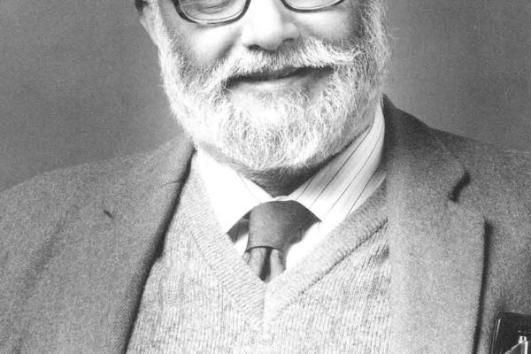 Abdus Salam, 1986 A portrait of Abdus Salam. He looks dignified, dressed in a wool jacket and a wool sweater underneath. He straightens his lapels as he smiles into the camera.