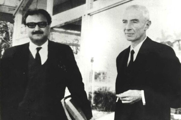 Salam and Oppenheimer, 1965 Salam, holding a stack of books and dressed in a suit, stands next to Oppenheimer outside a university building. Oppenheimer is holding a cigarette in his hand.